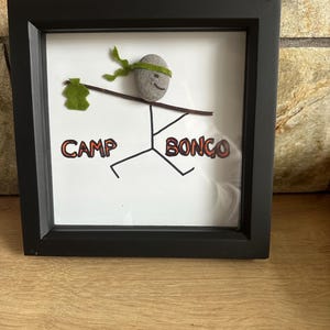 May include: A framed art piece with a black frame. The artwork features a stick figure with a stone head and a green headband, holding a green felt object. The words "CAMP BONGO" are printed below the figure.