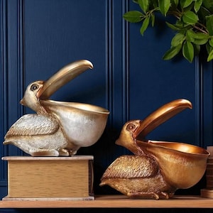 May include: Two decorative pelican sculptures in gold and silver tones, with open beaks and detailed feather textures. They are positioned on a wooden shelf against a dark blue background. These pelicans are decorative home decor items.