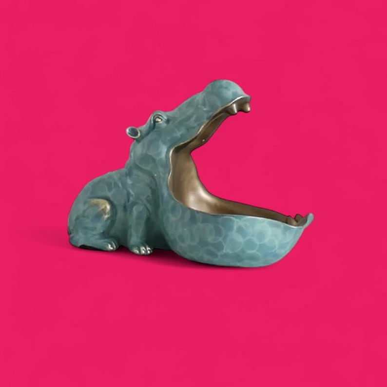 May include: A teal-colored hippo-shaped decorative bowl with an open mouth. The interior of the mouth is gold, and the bowl has a textured surface. The hippo is set against a bright pink background.
