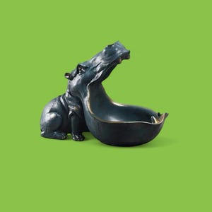 May include: A dark teal hippo-shaped decorative bowl with a bronze-colored rim. The hippo is in a sitting position with its mouth open, forming the bowl. The background is a solid green.