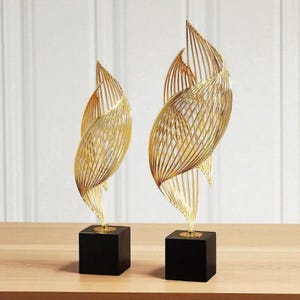 May include: Two gold-coloured metal sculptures with a spiral design, each mounted on a black square base. The sculptures have a delicate, openwork construction, creating a sense of lightness and elegance. The background is a light-coloured wall.