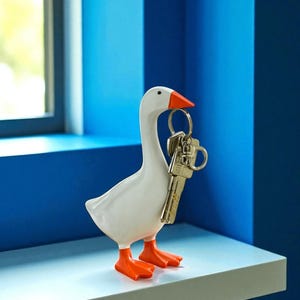 Op de afbeelding: Een witte ganzenvormige sleutelhouder met oranje voeten en snavel, die sleutels vasthoudt. De sleutelhouder staat op een lichtblauwe plank tegen een blauwe muur. Het is een decoratief item.