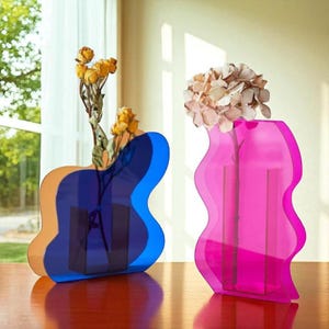 Op de afbeelding: Twee moderne vazen met golvende randen. Eén vaas heeft een gelaagd ontwerp van oranje, blauw en marine, met gele droogbloemen. De andere is felroze en bevat een beige bloemstuk. Beide vazen staan op een houten oppervlak.