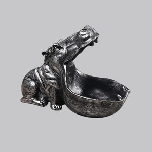 May include: A silver-colored hippo figurine with an open mouth forming a bowl. The hippo is in a seated position, with its mouth wide open, creating a functional bowl shape. The figurine has a distressed, metallic finish, adding a vintage look.