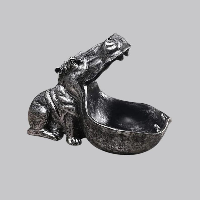 May include: A silver-colored hippo figurine with an open mouth forming a bowl. The hippo is in a seated position, with its mouth wide open, creating a functional bowl shape. The figurine has a distressed, metallic finish, adding a vintage look.