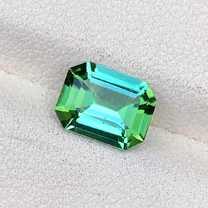 May include: A rectangular gemstone with a vibrant color gradient, transitioning from a bright green at the edges to a clear turquoise in the center. The gem is set against a white, textured surface, showcasing its facets and clarity.