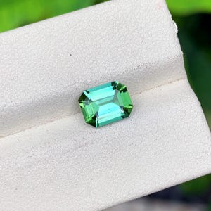 May include: A vibrant, emerald-cut gemstone with a striking green hue. The gem's facets reflect light, creating a brilliant display. The gemstone is set against a white surface, highlighting its color and clarity. This gemstone is perfect for jewelry.