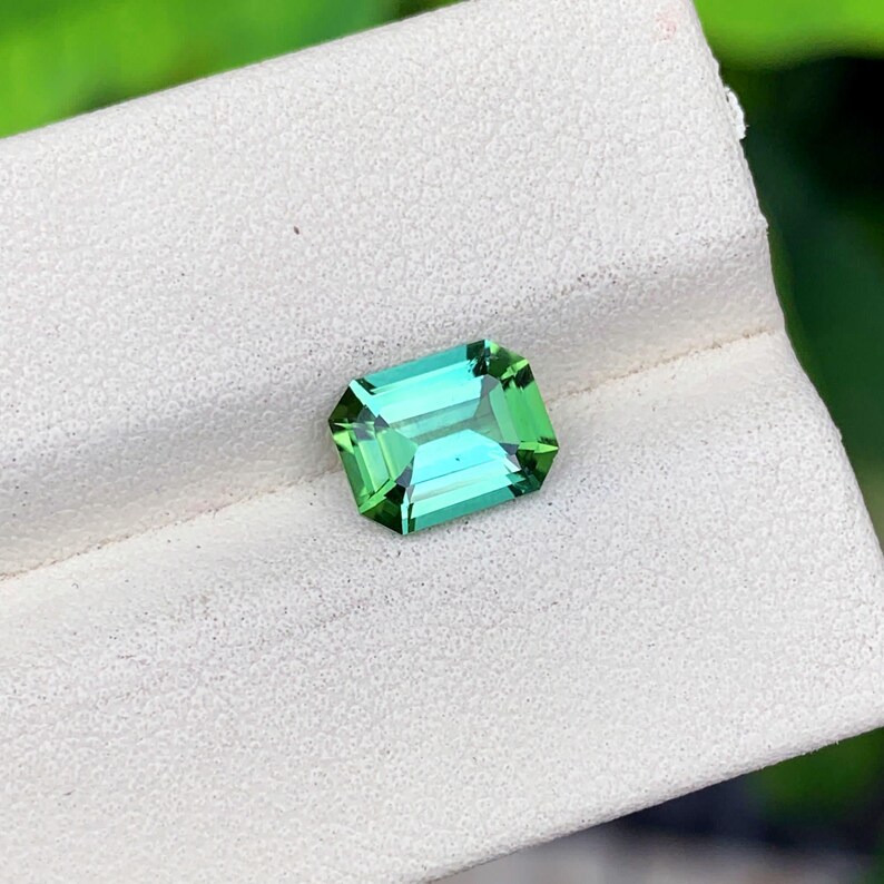 May include: A vibrant, emerald-cut gemstone with a striking green hue. The gem's facets reflect light, creating a brilliant display. The gemstone is set against a white surface, highlighting its color and clarity. This gemstone is perfect for jewelry.