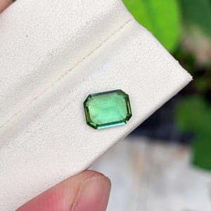 May include: A rectangular, emerald-cut gemstone with a gradient of green hues. The gem is set against a white background, showcasing its clarity and color. The gemstone's facets reflect light, highlighting its brilliance.