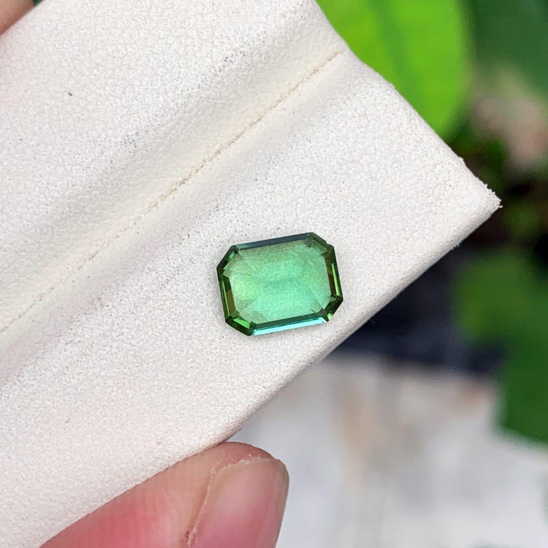 May include: A rectangular, emerald-cut gemstone with a gradient of green hues. The gem is set against a white background, showcasing its clarity and color. The gemstone's facets reflect light, highlighting its brilliance.