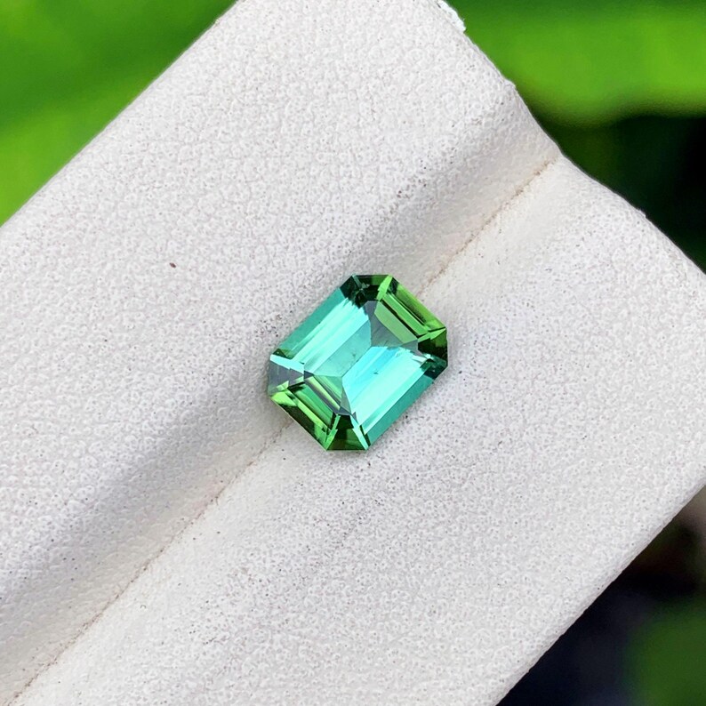 May include: A vibrant, emerald-cut gemstone with a striking green and teal color. The gem is set against a white background, showcasing its facets and clarity. The gemstone's rectangular shape and polished surface reflect light, highlighting its brilliance.