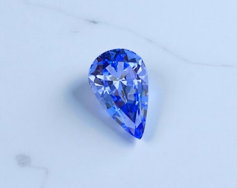 May include: A faceted, pear-shaped gemstone in a vibrant shade of blue. The gem's surface reflects light, showcasing its intricate cuts and angles. The stone is set against a white marble surface, highlighting its color and brilliance.