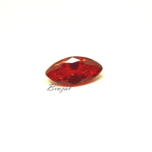 May include: A faceted, marquise-cut, red gemstone. The gem is a vibrant shade of red, with multiple facets that reflect light. The word "Binzal" is written in cursive on the lower left side of the gem. The gemstone is set against a white background.