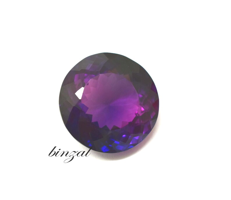 May include: A faceted, round gemstone with a gradient of purple and blue hues. The gem has a polished surface and is set against a white background. The word "binzal" is written in cursive on the lower left.