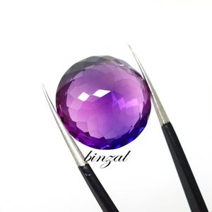 May include: A faceted, round amethyst gemstone with a gradient of purple hues, held by tweezers against a white background. The word "binzal" is written in cursive on the lower right side of the image.