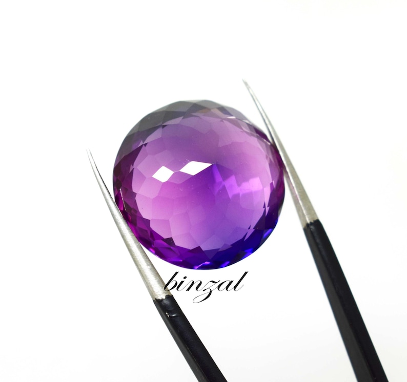 May include: A faceted, round amethyst gemstone with a gradient of purple hues, held by tweezers against a white background. The word "binzal" is written in cursive on the lower right side of the image.