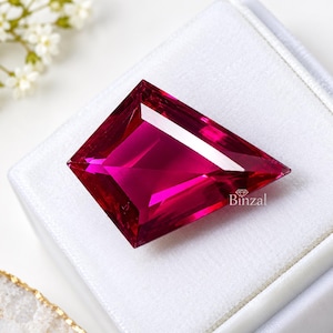 Op de afbeelding: Een vliegervormige, diep magenta edelsteen met een gefacetteerd oppervlak en scherpe randen. De steen heeft een briljante, reflecterende helderheid en ligt op een wit fluwelen displaykussen. Het woord Binzal staat in kleine letters op de steen gedrukt.