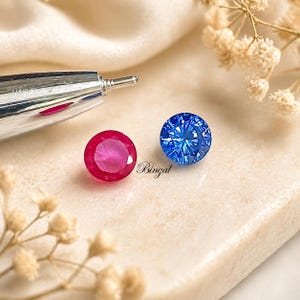 Peut inclure: Deux pierres précieuses rondes : un rubis et un saphir. Le rubis est d'un rouge rosé profond, tandis que le saphir est d'un bleu vif et facetté. Les pierres précieuses sont présentées sur une surface claire avec le mot "Bingal" en cursive.