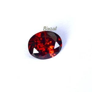 May include: A faceted, oval-shaped, deep red gemstone. The gem has a brilliant, fiery appearance with multiple facets reflecting light. The word "Binzal" is printed in a light green color on the upper right side of the image.