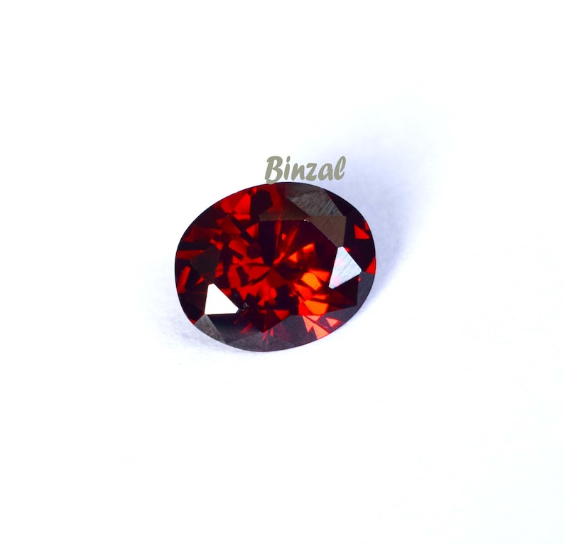 May include: A faceted, oval-shaped, deep red gemstone. The gem has a brilliant, fiery appearance with multiple facets reflecting light. The word "Binzal" is printed in a light green color on the upper right side of the image.