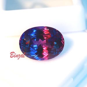 May include: An oval-cut gemstone with a vibrant display of colors, including blue, purple, and red hues. The gemstone is faceted, creating a sparkling effect. The word "Binzal" is written in red cursive on the lower left.