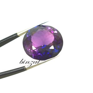 May include: A faceted, oval-cut gemstone with a deep purple hue, transitioning to blue in certain areas. The gem is held by a pair of tweezers against a white background. The word "binzal" is printed on the image.