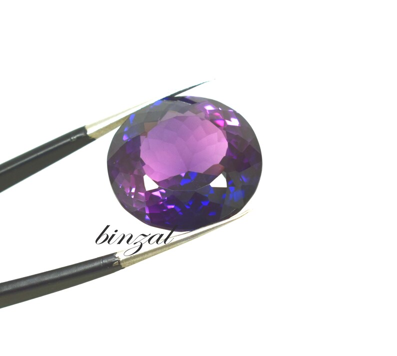 May include: A faceted, oval-cut gemstone with a deep purple hue, transitioning to blue in certain areas. The gem is held by a pair of tweezers against a white background. The word "binzal" is printed on the image.