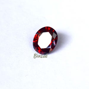May include: A faceted, oval-shaped, deep red gemstone. The gem has a polished surface with multiple facets that reflect light. The word "Binzal" is printed in light grey at the bottom of the image. The gemstone is set against a white background.
