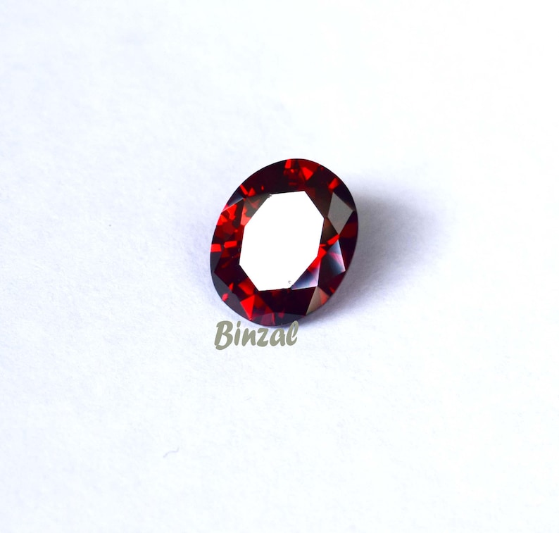 May include: A faceted, oval-shaped, deep red gemstone. The gem has a polished surface with multiple facets that reflect light. The word "Binzal" is printed in light grey at the bottom of the image. The gemstone is set against a white background.