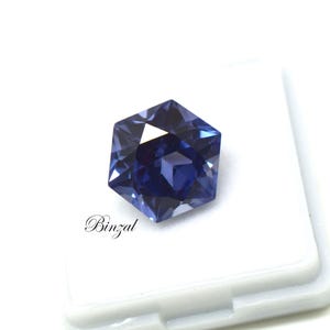 Puede incluir: Una piedra preciosa hexagonal facetada con un tono azul y violeta intenso, que muestra cortes geom&eacute;tricos n&iacute;tidos y reflejos de luz. La piedra est&aacute; colocada sobre una base blanca con el texto Binzal impreso en negro en letra cursiva.