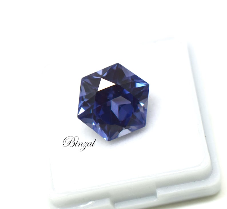 Puede incluir: Una piedra preciosa hexagonal facetada con un tono azul y violeta intenso, que muestra cortes geom&eacute;tricos n&iacute;tidos y reflejos de luz. La piedra est&aacute; colocada sobre una base blanca con el texto Binzal impreso en negro en letra cursiva.