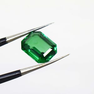 May include: A vibrant, emerald-green gemstone cut in a rectangular, step-faceted shape. The stone is held by a pair of thin, silver-toned metal tweezers with black handles, showcasing the clarity and deep green color of the faceted gemstone.