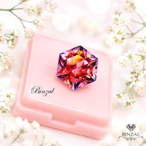 Puede incluir: Una piedra preciosa hexagonal facetada con un degradado vibrante de tonos rojos, naranjas y morados, sobre un estuche cuadrado de color rosa claro. El estuche presenta el texto Binzal en letra cursiva negra y un logo con el texto BINZAL.