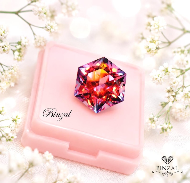 Puede incluir: Una piedra preciosa hexagonal facetada con un degradado vibrante de tonos rojos, naranjas y morados, sobre un estuche cuadrado de color rosa claro. El estuche presenta el texto Binzal en letra cursiva negra y un logo con el texto BINZAL.