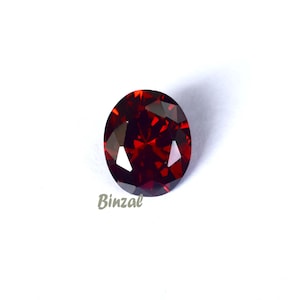 May include: A faceted, oval-shaped, deep red gemstone. The gem is set against a white background, with the word "Binzal" in a light gray script in the lower left corner. The gem's facets reflect light, creating a sparkling effect.