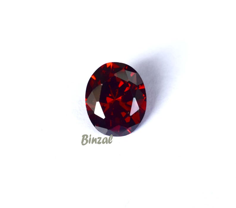 May include: A faceted, oval-shaped, deep red gemstone. The gem is set against a white background, with the word "Binzal" in a light gray script in the lower left corner. The gem's facets reflect light, creating a sparkling effect.