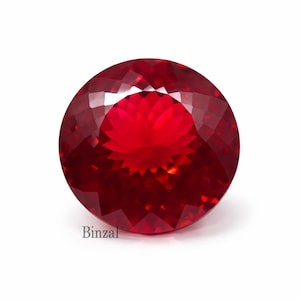 May include: A faceted, round, deep red gemstone. The gem has a brilliant cut with many facets, reflecting light. The color is a rich, saturated red, with a slight variation in tone across the surface. The word "Binzal" is printed on the bottom left.
