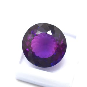 May include: A round, faceted gemstone with a deep purple hue, transitioning to lighter shades in the center. The gem is set against a white background, showcasing its brilliant cut and color variations. This gemstone is ideal for jewelry.