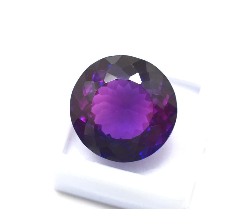 May include: A round, faceted gemstone with a deep purple hue, transitioning to lighter shades in the center. The gem is set against a white background, showcasing its brilliant cut and color variations. This gemstone is ideal for jewelry.