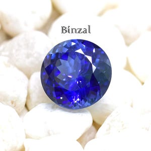May include: A round, deep blue gemstone with multiple facets, likely a sapphire, rests on white pebbles. The word "Binzal" is written above the gemstone. The gemstone is a striking shade of blue.