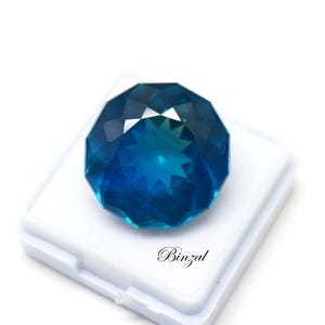 May include: A round, faceted, deep blue gemstone with a star-shaped pattern at its center. The stone is displayed on a white square base. The text "Binzal" is printed in black script on the white surface below the gemstone.
