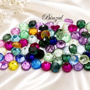 Può includere: Una collezione di gemme sfaccettate in vari colori, tra cui rosa, verde, blu e giallo, sparse su una superficie di raso bianco. La parola "Binzal" è scritta in corsivo. Piccoli fiori bianchi nell'angolo in alto a sinistra.