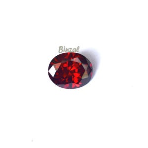 May include: A faceted, oval-shaped, deep red gemstone. The gem has a brilliant cut, reflecting light. The background is plain white. The word "Binzal" is printed in light green in the upper left corner.