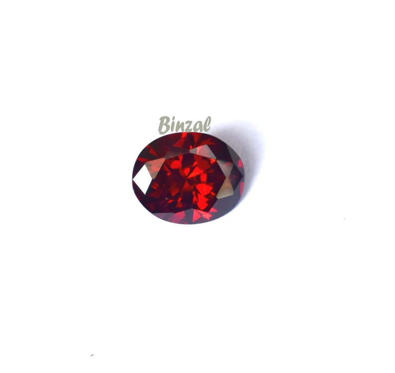 May include: A faceted, oval-shaped, deep red gemstone. The gem has a brilliant cut, reflecting light. The background is plain white. The word "Binzal" is printed in light green in the upper left corner.