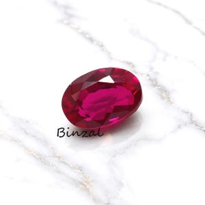 May include: A faceted, oval-shaped ruby gemstone with a deep red hue. The gem is set against a white marble surface. The word "Binzal" is written in cursive script near the bottom of the image.