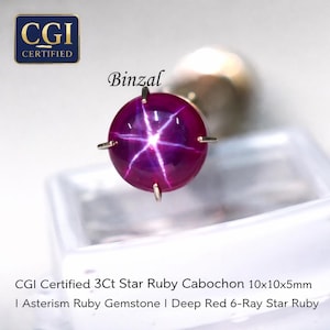 May include: A deep red star ruby cabochon gemstone with a six-ray star pattern. The gem is round, approximately 0.4 inches in diameter, and set with small metal prongs. The image includes the text "CGI Certified" and "Binzal".