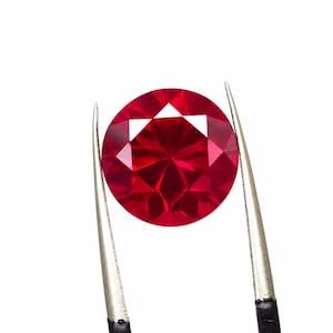 Puede incluir: Una gema de rubí redonda y facetada sostenida por pinzas plateadas sobre un fondo blanco. El rubí es de un color rojo intenso y vibrante, con múltiples facetas que reflejan la luz. Las pinzas tienen mangos negros.