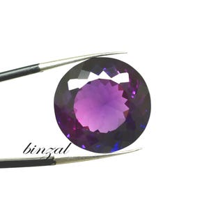 May include: A faceted, round amethyst gemstone, held by tweezers against a white background. The gem displays a gradient of purple hues, from deep violet to lighter shades in the center. The word "binzal" is printed on the lower left.