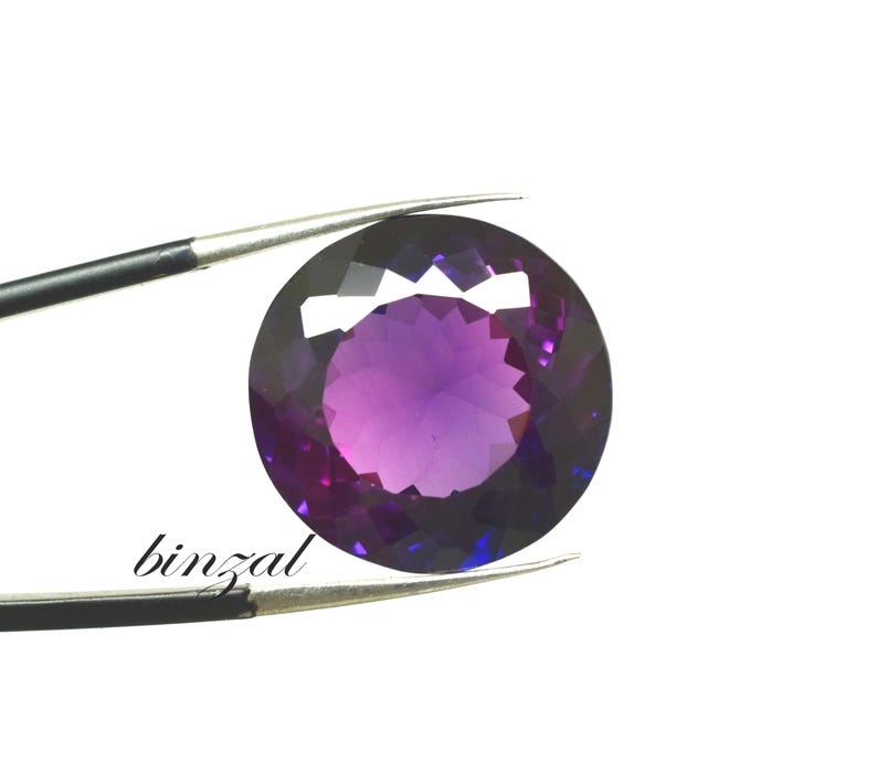 May include: A faceted, round amethyst gemstone, held by tweezers against a white background. The gem displays a gradient of purple hues, from deep violet to lighter shades in the center. The word "binzal" is printed on the lower left.
