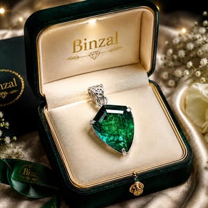 Könnte beinhalten: Ein schildförmiger grüner Edelsteinanhänger mit einer silbernen Filigranöse, präsentiert in einer dunkelgrünen Samtschmuckschatulle mit dem goldenen Schriftzug Binzal auf dem cremefarbenen Innendeckel und einer passenden Box.